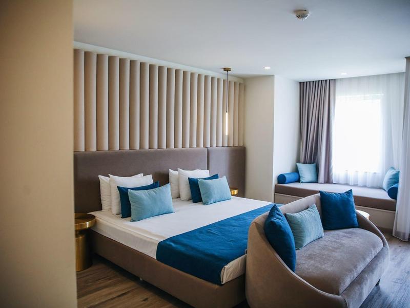 Moderne hotelkamer met tweepersoonsbed, blauwe kussens en zithoek bij het raam.