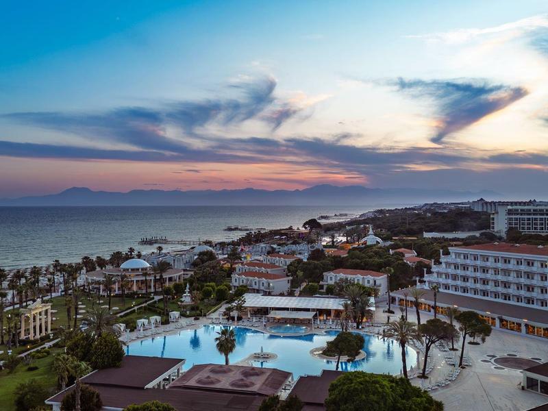 Uitzicht op een resort met zwembad, palmbomen en zee bij zonsondergang.