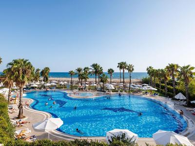 Großer blauer Pool mit Palmen, weißen Liegen und Blick auf das Meer unter klarem Himmel.