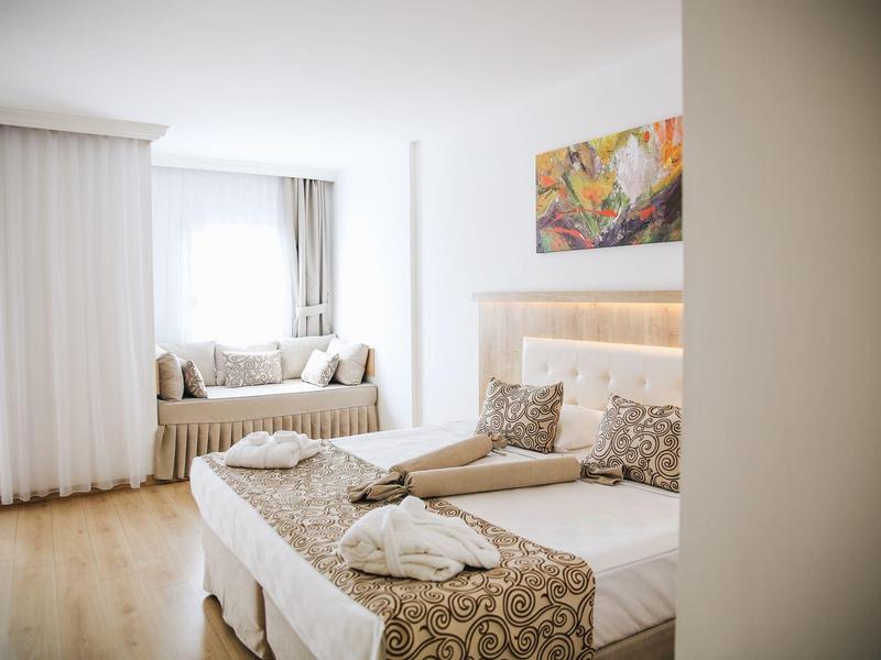 Camera d'hotel luminosa con letto matrimoniale, cuscini decorativi, seduta finestra e quadro colorato.