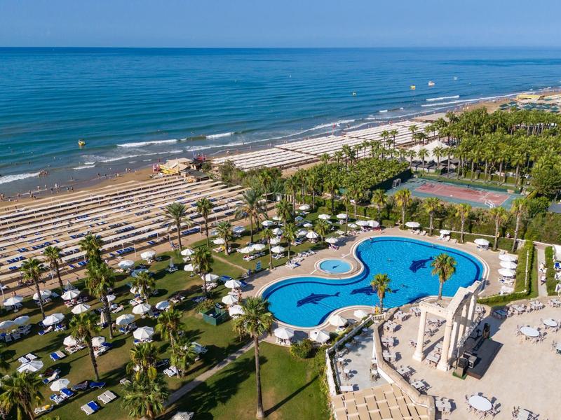 Veduta aerea di un resort sulla spiaggia con una grande piscina, lettini, palme e il mare sullo sfondo.