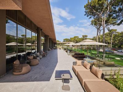 Area esterna moderna dell'hotel con posti a sedere, circondata da alberi sotto un cielo azzurro.