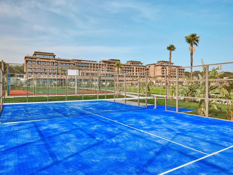 Campo da padel blu con edifici dell'hotel e palme sullo sfondo sotto un cielo azzurro.