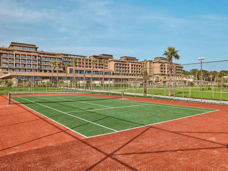 Un campo da tennis con superficie rossa davanti a un grande complesso alberghiero sotto un cielo blu.