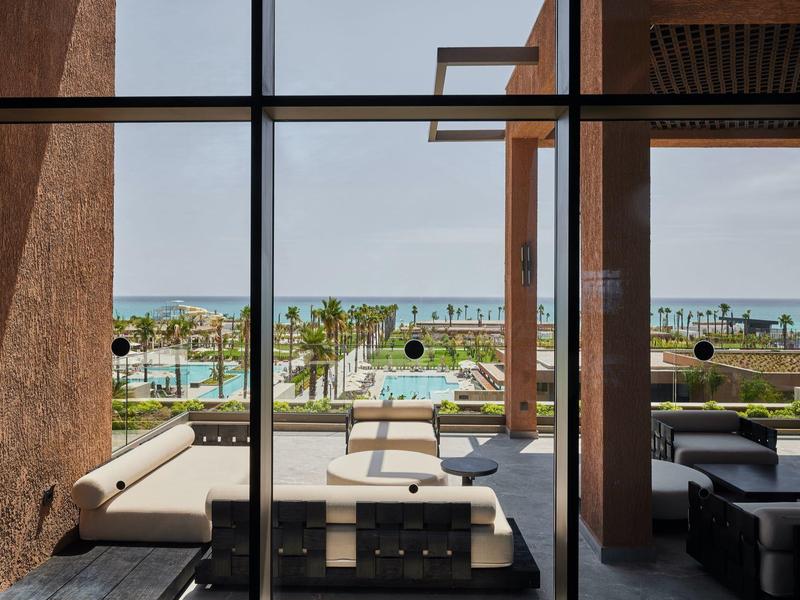 Vista attraverso finestre di vetro su terrazza con divani e piscina, mare sullo sfondo.