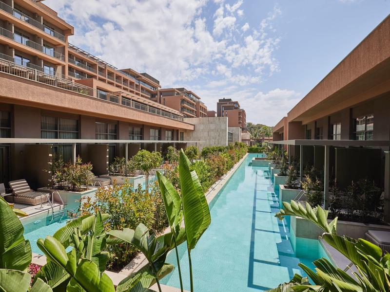 Cortile moderno di un hotel con piscina e lettini, circondato da alberi e edifici.