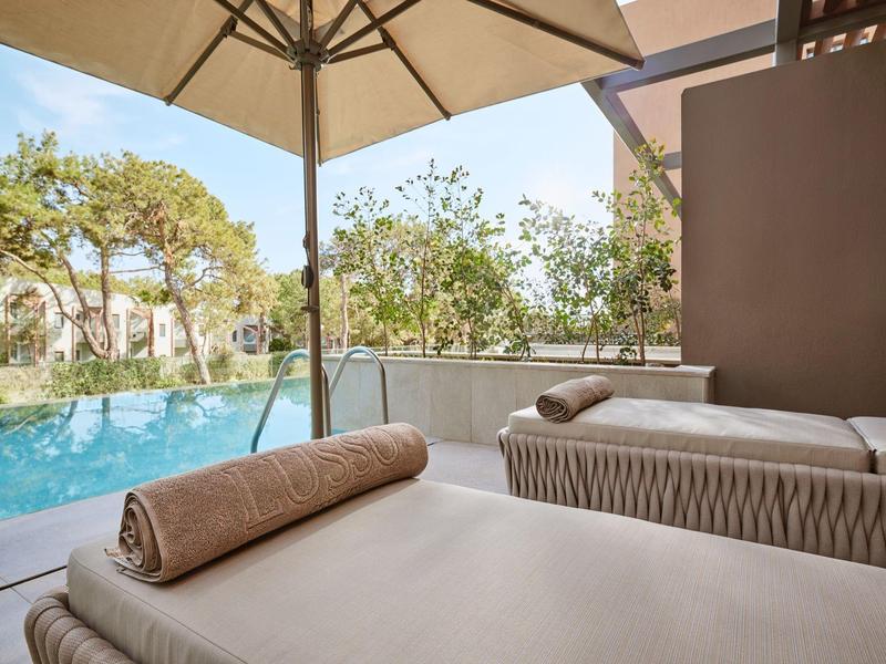 Lettini con asciugamani sotto ombrellone vicino a piscina e alberi.