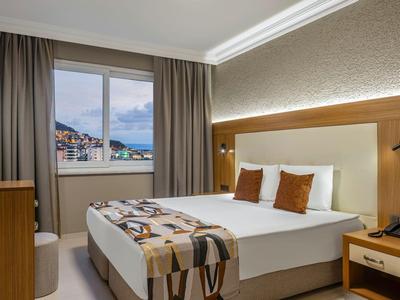 Helles Hotelzimmer mit Doppelbett, Nachttisch, Fensterblick auf Stadt und Bergen.
