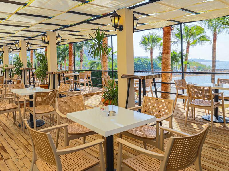 Otevřená terasa restaurace se stoly, židlemi a výhledem na moře pod palmami.