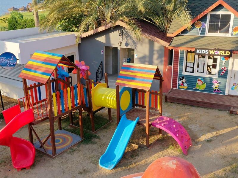 Barevné dětské hřiště se skluzavkami a tunelem před rekreačním domem pod palmami.