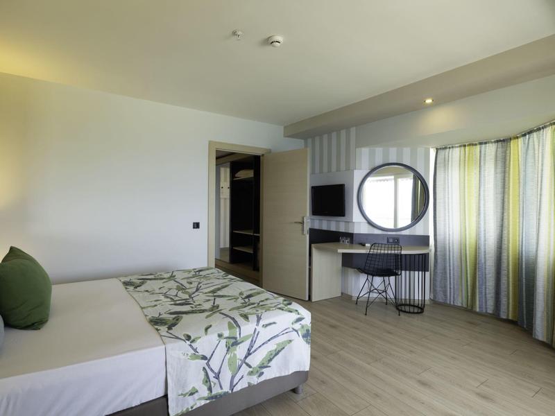 Moderní hotelový pokoj s manželskou postelí, psacím stolem, židlí a velkými okny se závěsy.