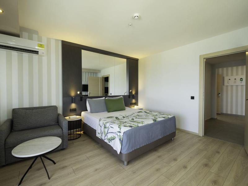 Moderní hotelový pokoj s manželskou postelí, křesly, zrcadlem a skříní v současném stylu.