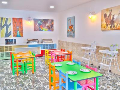 Barevná dětská herna s malými stoly, jasnými židlemi a dětskými knihami na stěně.