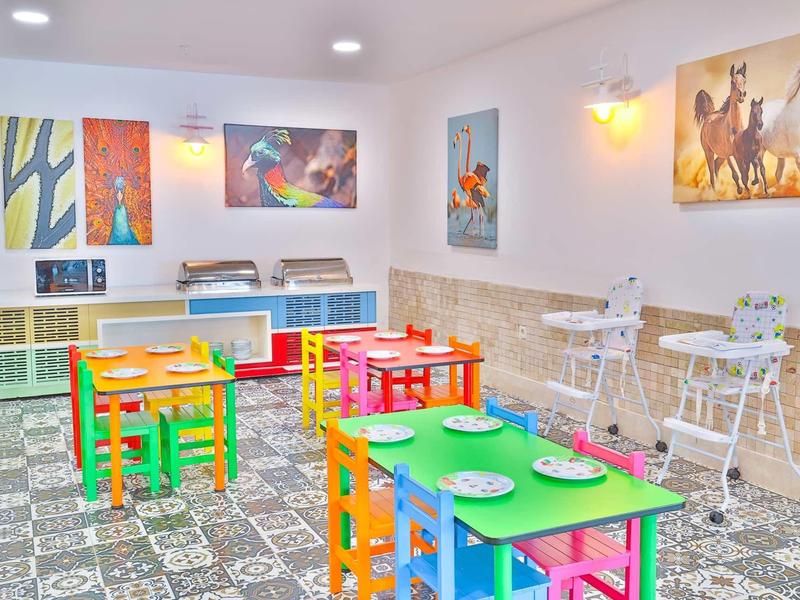 Barevná dětská herna s malými stoly, jasnými židlemi a dětskými knihami na stěně.