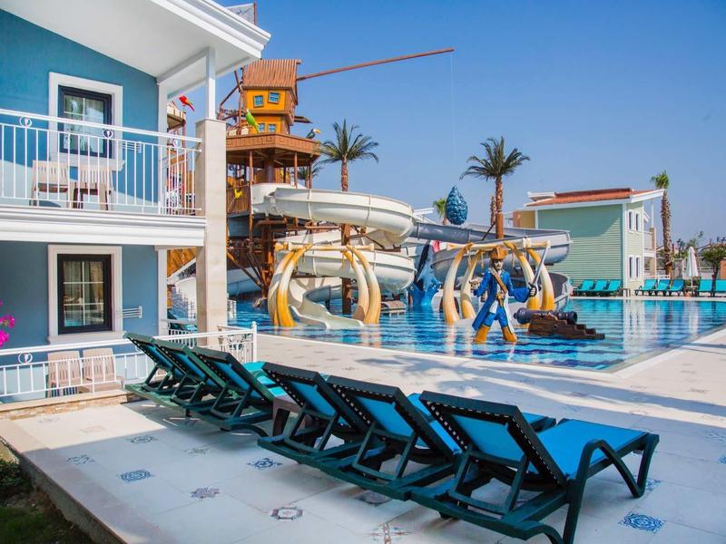 Bazénová oblast s lehátky a vodními skluzavkami vedle vícepodlažního hotelu pod modrou oblohou.
