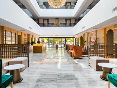 Heller Hotel-Lobbybereich mit Marmorboden, grünen Sesseln, Holzpanelen und großem Empfangstresen.