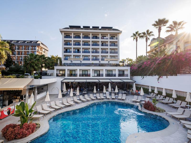 Hotel with large pool, surrounded by lounge chairs, palm trees, and modern architecture in sunlight.