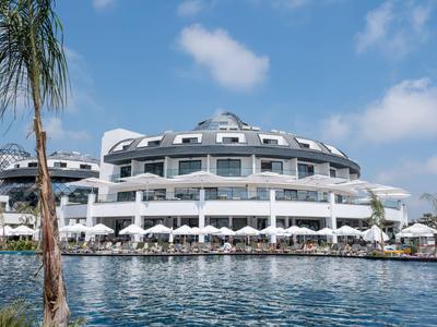 Modernes rundes Hotel am Wasser mit Terrassen und Sonnenschirmen unter blauem Himmel.