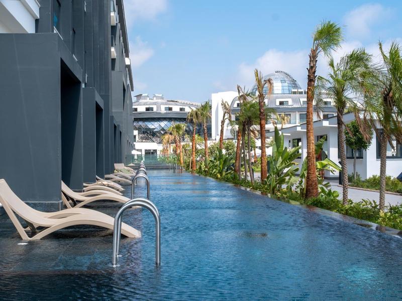 Moderner Pool mit Liegen neben einem Hotel und Palmen unter blauem Himmel.