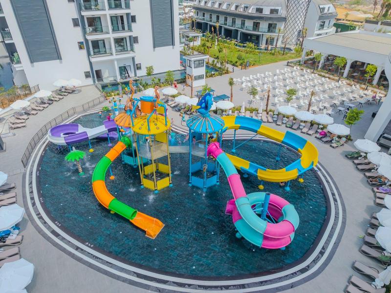 Bunter Wasserspielbereich mit Rutschen und Wasserfontänen in einem Hotelpool, umgeben von Liegestühlen.