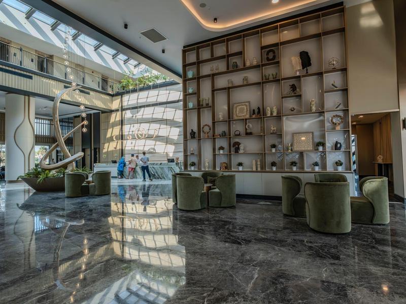 Hotel-Lobby mit Marmorboden, grünen Sesseln und großer Wand mit dekorativen Objekten.