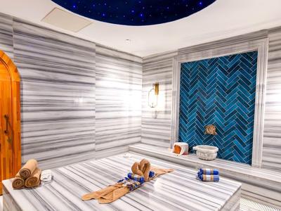 Helles Hamam mit weißen Marmorwänden, Holz-Tür, blauer Kachelwand und Wasserbecken.