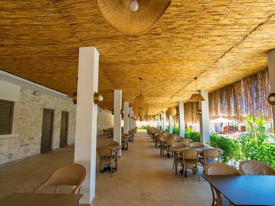 Zastřešená venkovní plocha s stoly a židlemi, obklopená rostlinami a přirozeným světlem.