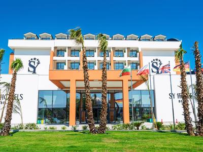 Moderní hotelová budova se skleněnou fasádou a palmami na zeleném trávníku pod jasnou oblohou.