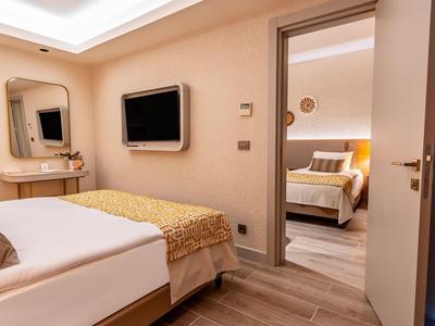 Pohodlný hotelový pokoj s postelí, televizí na stěně, toaletním stolkem a výhledem do druhého pokoje.