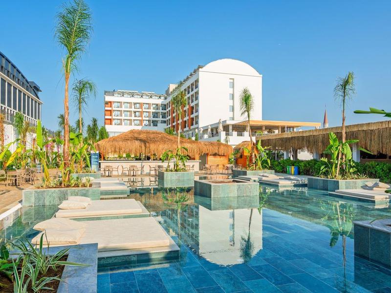 Moderner Hotelpool mit Palmen, Liegen und klar blauem Wasser unter hellem Himmel.