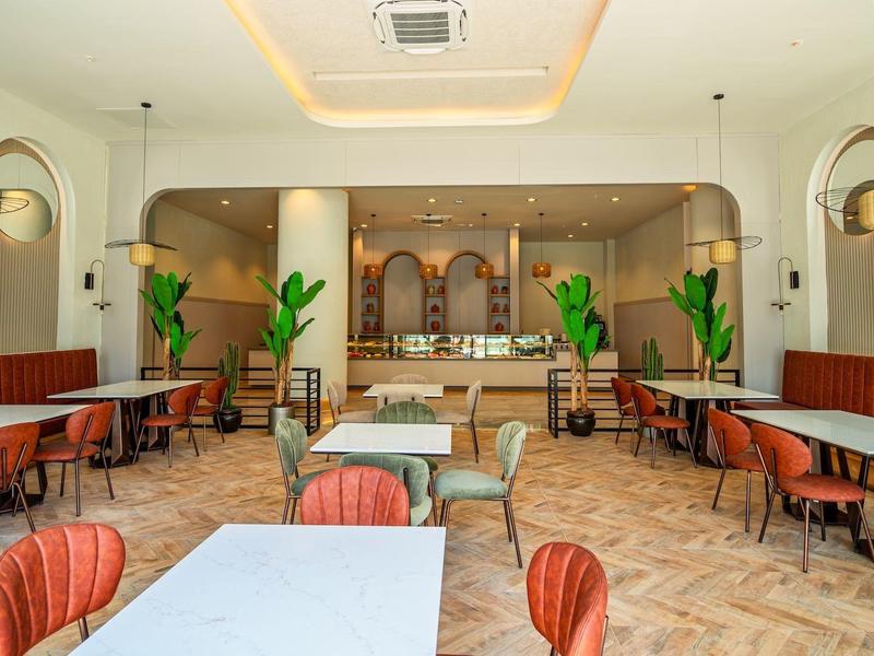 Heller, moderner Gastraum mit roten Stühlen, weißen Tischen, Pflanzen und großen Bogenfenstern.