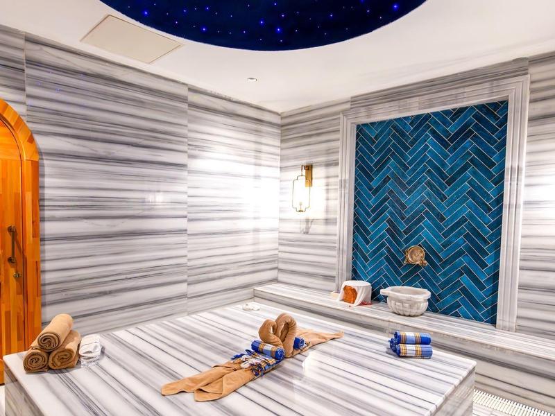 Helles Hamam mit weißen Marmorwänden, Holz-Tür, blauer Kachelwand und Wasserbecken.