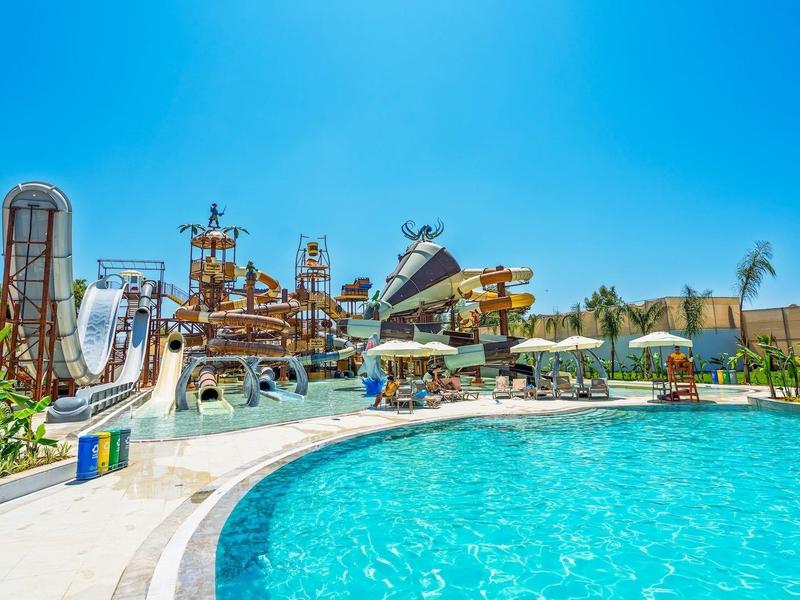 Wasserpark mit großem Pool, Rutschen, Wasserspielen und Palmen unter klarem blauem Himmel.