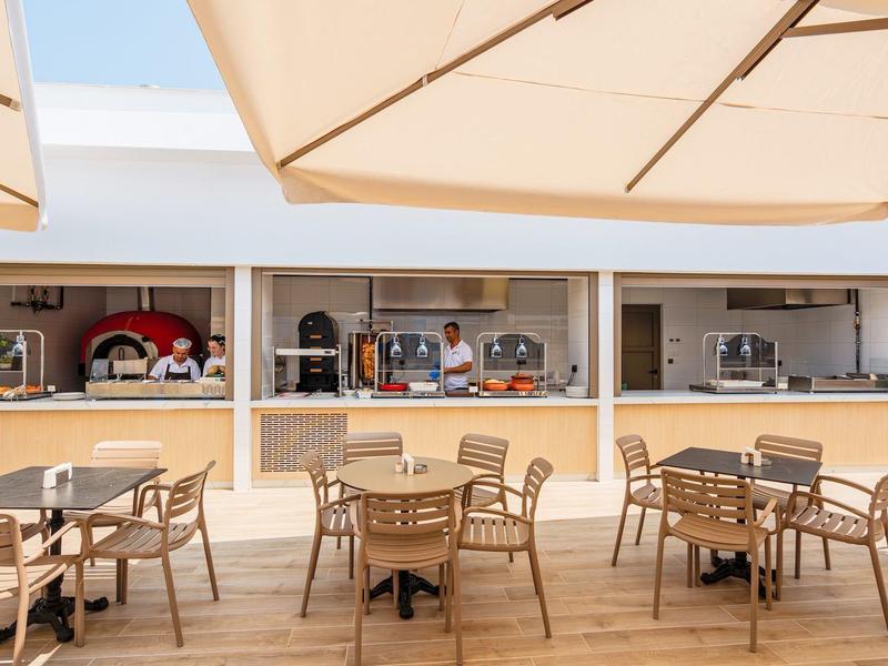 Terrasse mit Holztischen, Stühlen und großen Sonnenschirmen vor einem Café mit offenem Schalter.