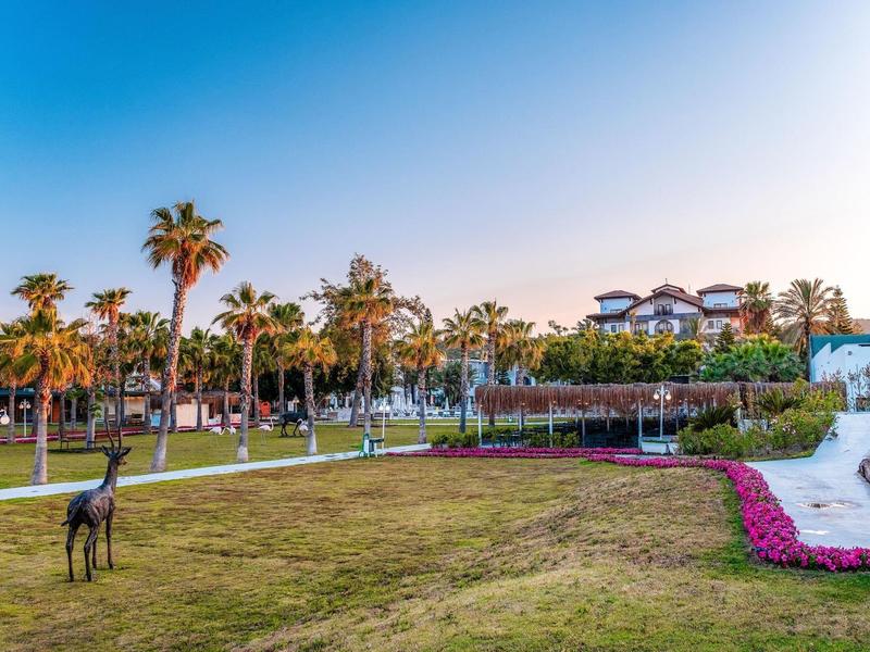 Espace herbeux avec sculptures, massifs de fleurs et palmiers devant un bâtiment au coucher du soleil.