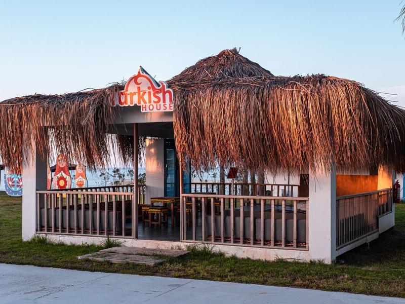 Petit restaurant avec toit de chaume et terrasse au bord d'un lac sous un ciel clair.