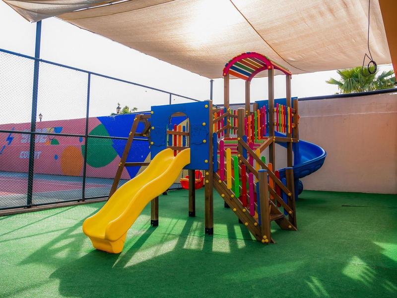 Aire de jeux pour enfants avec toboggan jaune et structure d'escalade sous une toile d'ombrage.