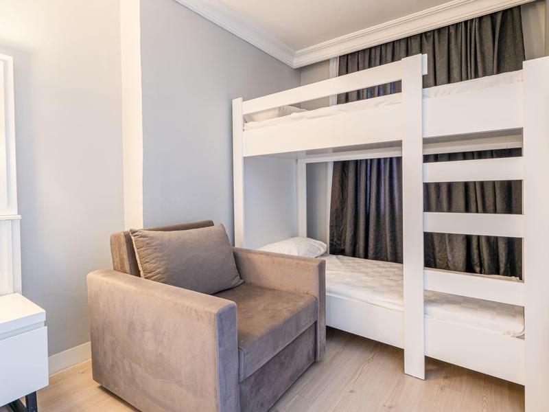 Petite chambre avec fauteuil gris et lit superposé blanc, lumière vive de la fenêtre et rideaux.