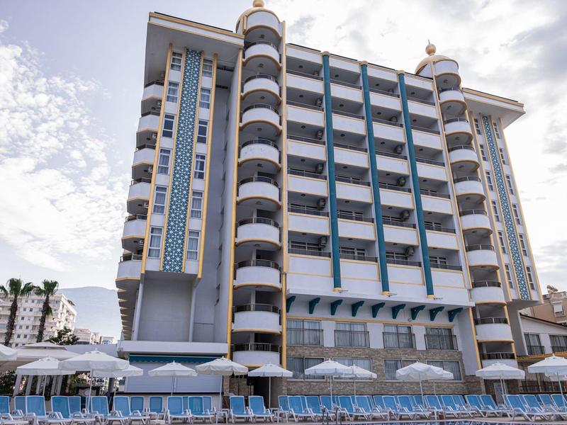 Hôtel à plusieurs étages avec balcons et parasols bleus sur la plage sous un ciel nuageux.