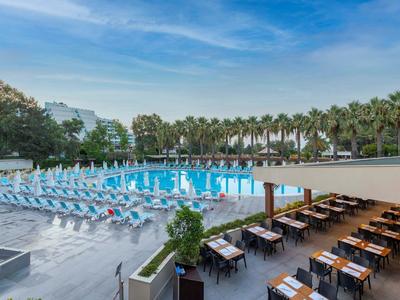 Grande piscina dell'hotel con lettini e area esterna ombreggiata con tavoli e sedie.