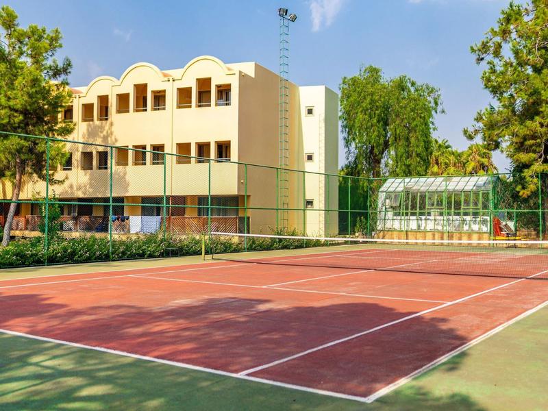 Pista de tenis vacía junto a un edificio de hotel con árboles y invernadero al fondo.