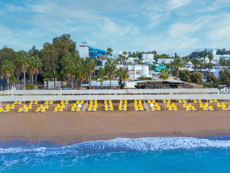 Playa con tumbonas amarillas, sombrillas, mar azul y edificios blancos sobre colinas.
