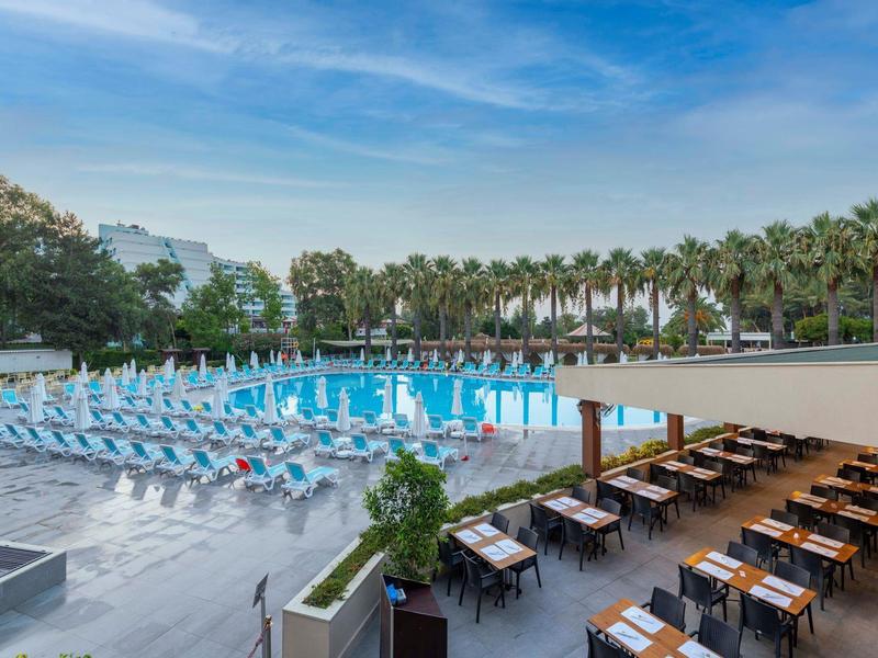 Gran piscina de hotel con muchas tumbonas y restaurante exterior techado bajo un cielo despejado.