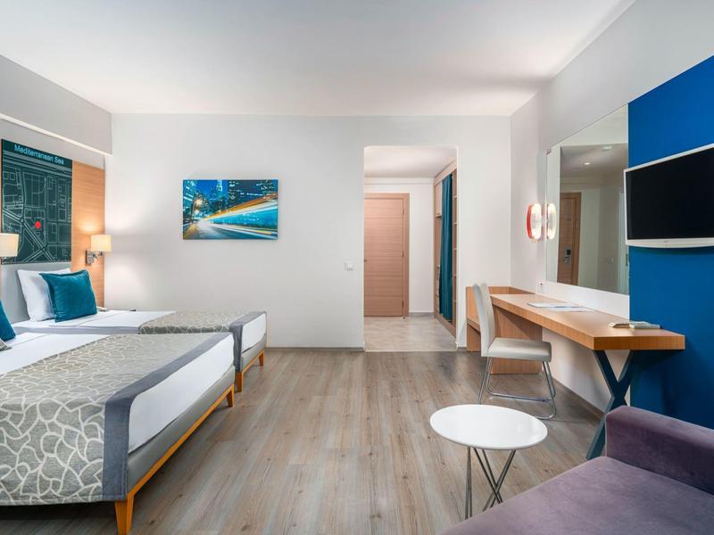 Camera d'hotel moderna con due letti, pavimento in legno, scrivania e TV su una parete blu.