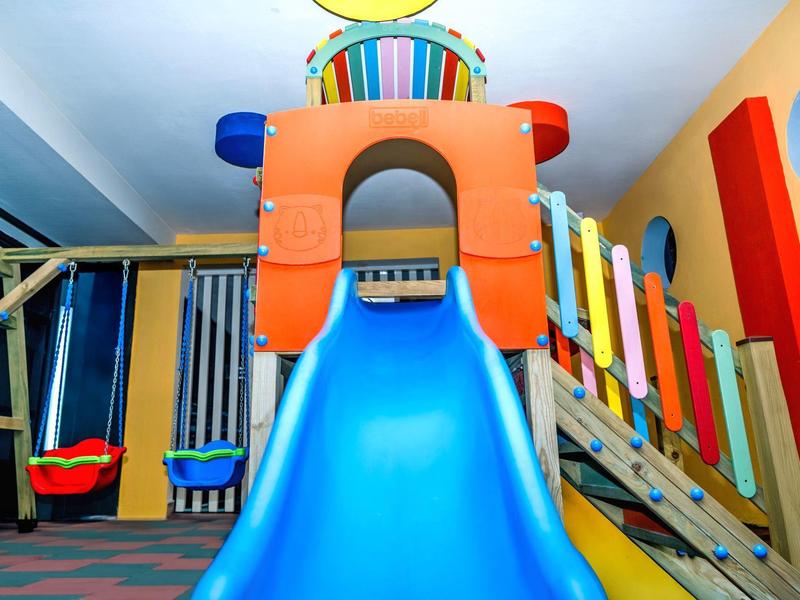 Parco giochi colorato al coperto con scivolo e altalene in una stanza luminosa.