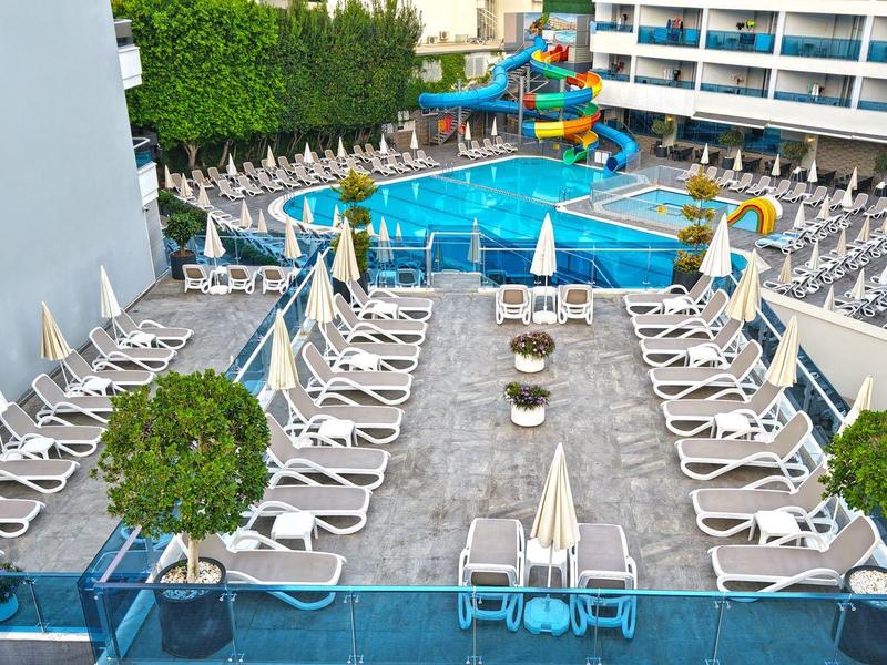 Vista di una piscina d'hotel con ombrelloni e lettini disposti in file, circondata da un edificio su più piani.