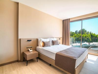 Helles Hotelzimmer mit großem Bett, Nachtisch und Blick auf Pool und Bäume durch großes Fenster.