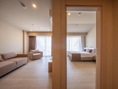Helles Hotelzimmer mit getrenntem Wohnbereich und Schlafzimmer, große Fenster mit Vorhängen.