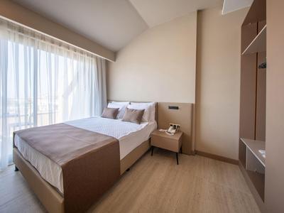 Schlicht eingerichtetes Hotelzimmer mit Doppelbett, Nachttisch und großem Fenster mit Gardinen.