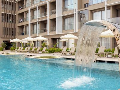 Moderner Hotelpool mit Wasserfall, Liegestühlen und Sonnenschirmen vor dem Gebäude.